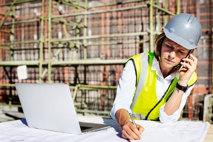 Female construction architect takes phone call beside laptop and drawings
