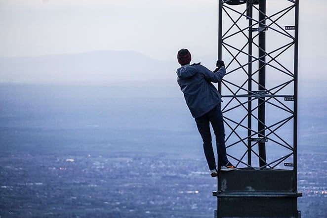 A man hangs from cell tower observing an urban scene