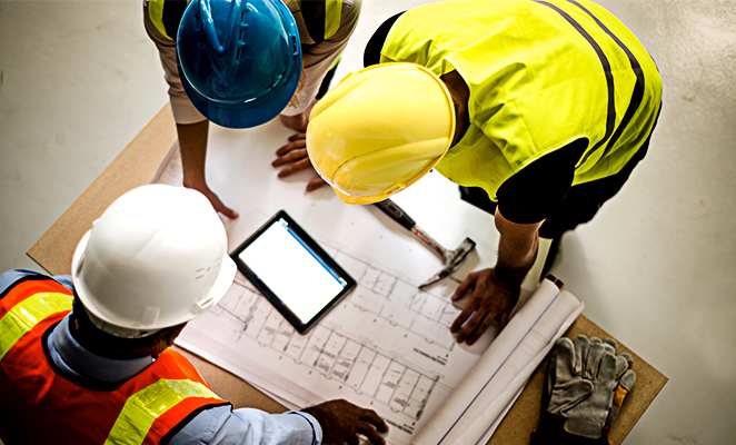 Construction workers wearing hard hats and high visibility vests huddle around building plans and tablet reader