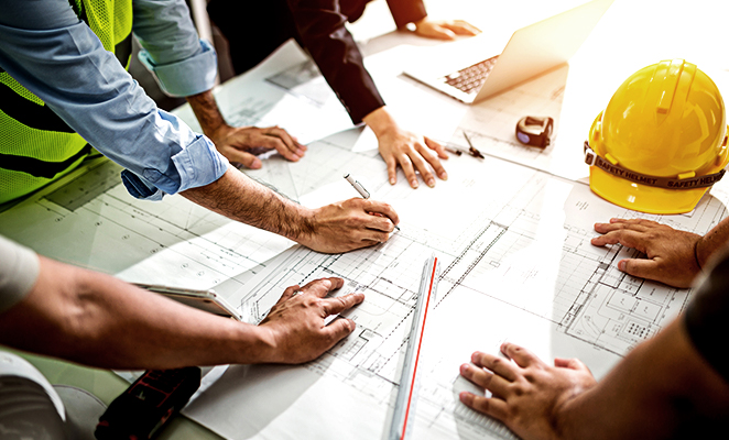 A group of contractors sketch construction plans over desk beside yellow hard hat