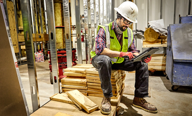 A contractor wearing high visibility vest and hard hat in build site sits on top of wooden planks while viewing iPad 