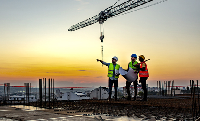 Three construction workers in high visibility vests and hard hats scan jobsite with one in the middle holding build plans