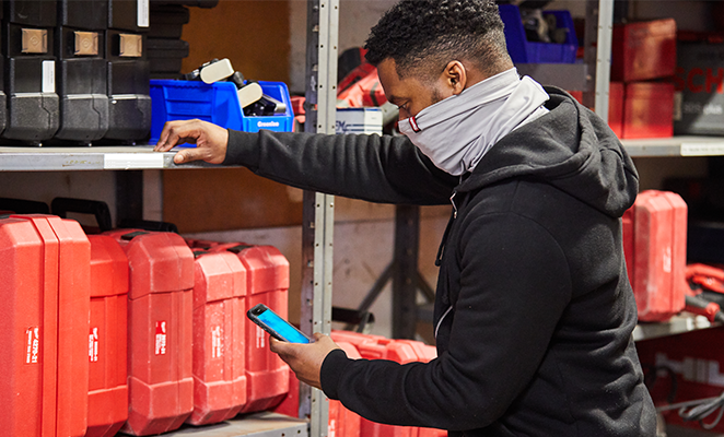 A tool room manager wearing gray Milwaukee gaiter looks at his smartphone next to shelving full of tools in red boxes