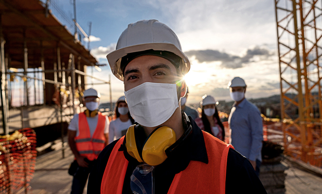 A masked contractor in a high-visibility vest leads a group of male and female workers on site