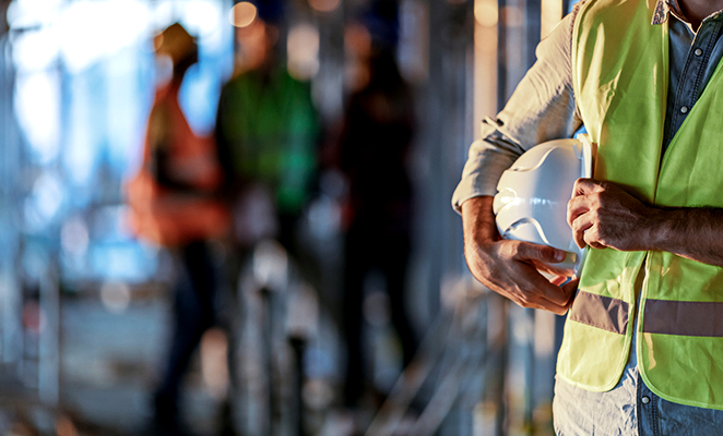 A construction worker contemplatively stands holding white hard hat while an out-of-focus trio of workers behind him talk