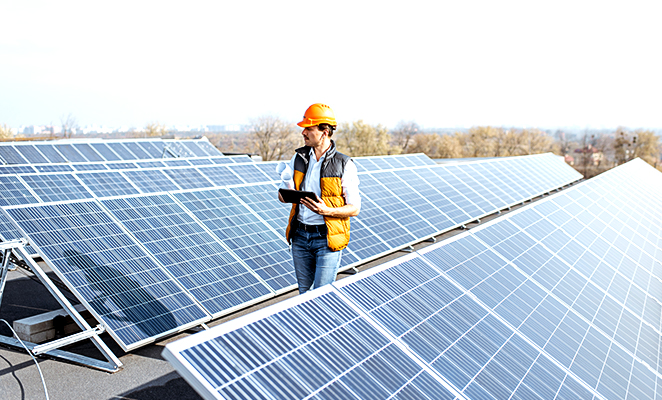 On building roof, a young solar photovoltaic installer with iPad inspects solar panels