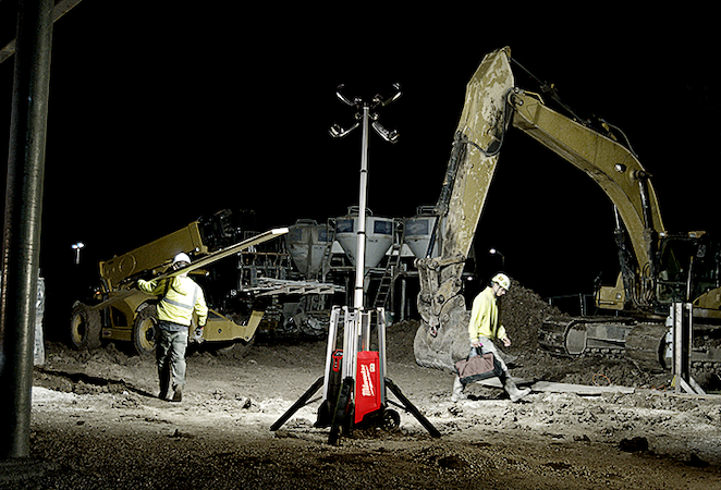 A Milwaukee light tower illuminates construction site with two workers walking opposite directions