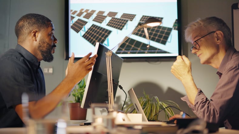 Two colleagues have a conversation with solar panels displayed on a wall-mounted monitor