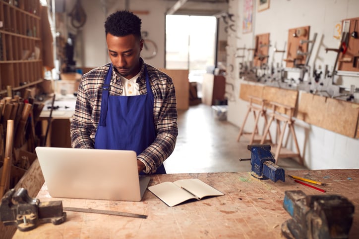 A young carpenter in woodshed uses laptop to complete customer order