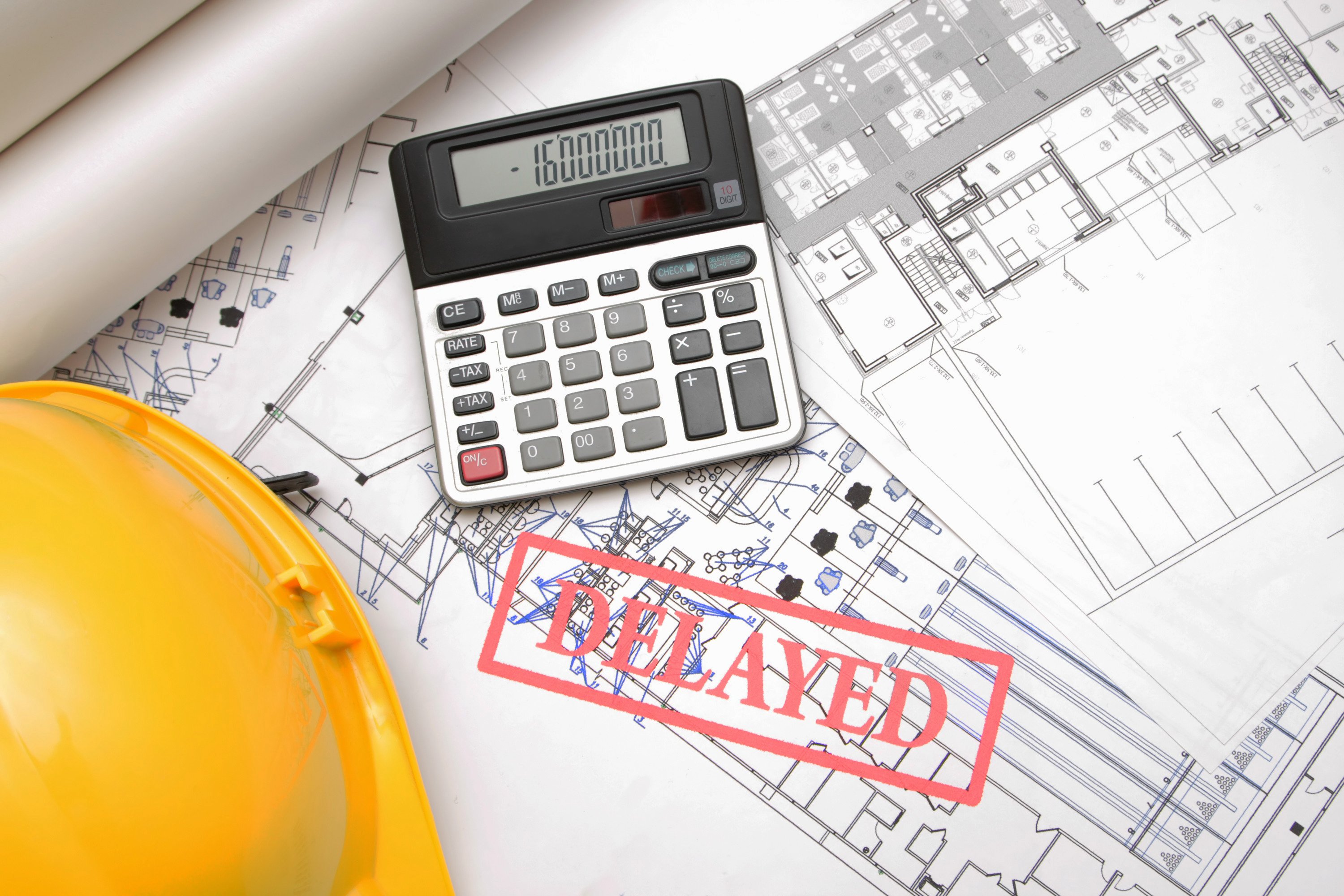 Project plans strewn across contractor's desk beside calculator displaying negative calculations. Word “delayed” overlain  