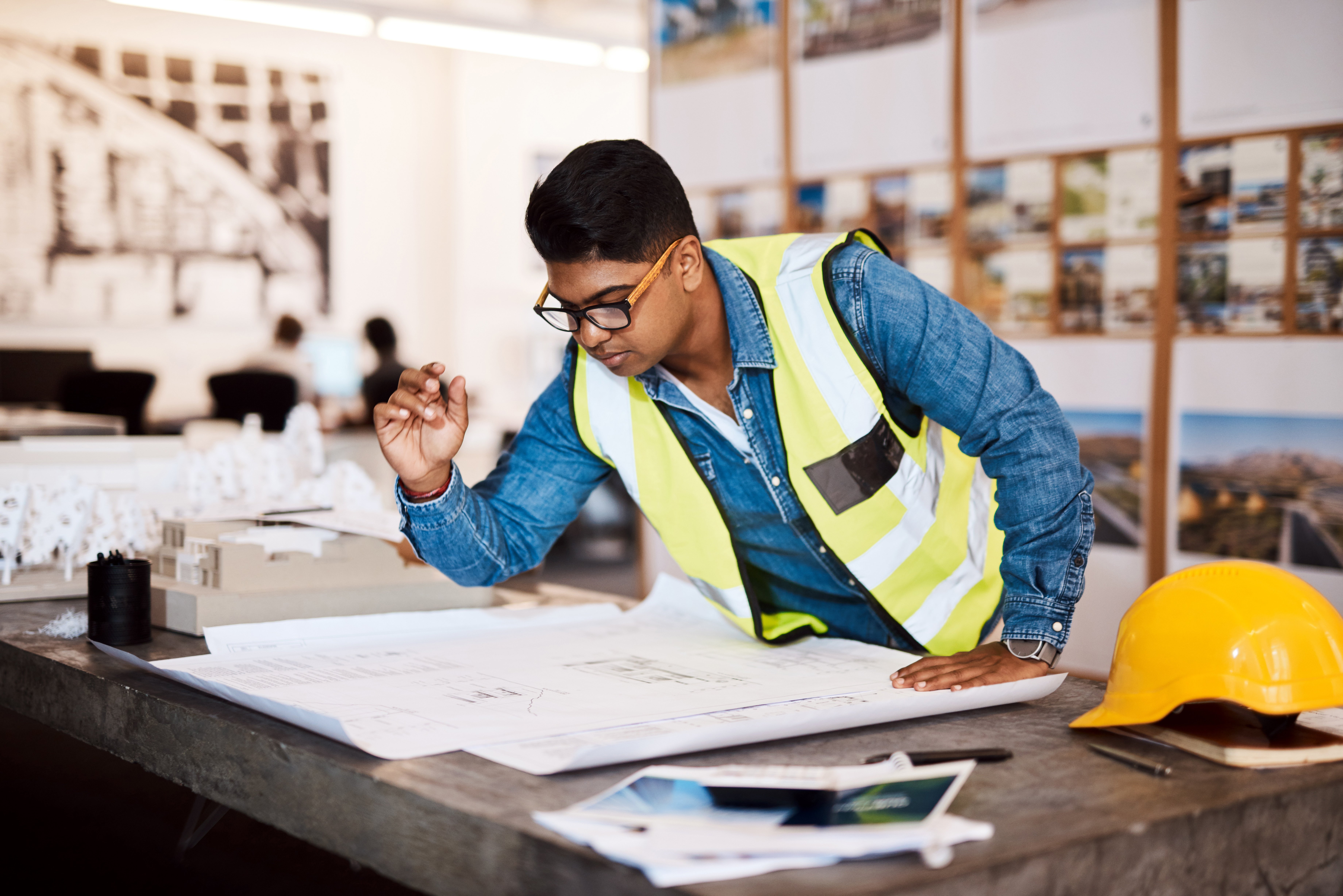 A construction project manager at desk wearing high-vis jacket, beside yellow hard hat, makes up architectural plans