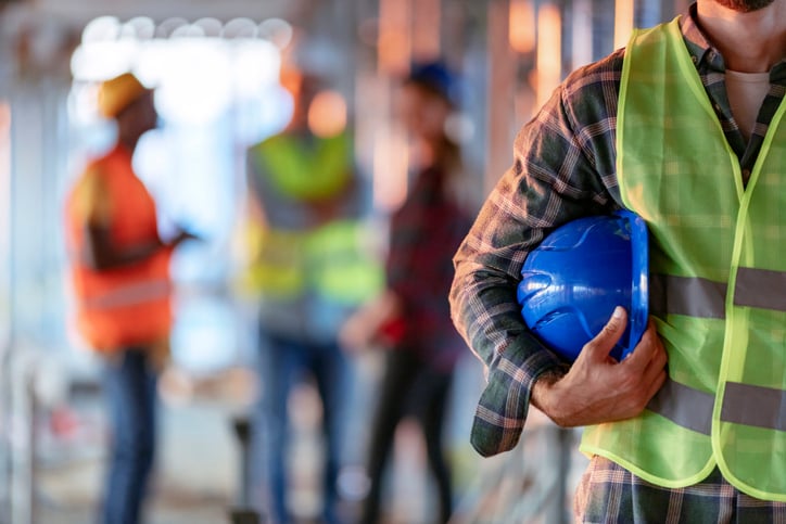 A construction worker in high-visibility vest holds blue hardhat beside 3 workers talking in the background