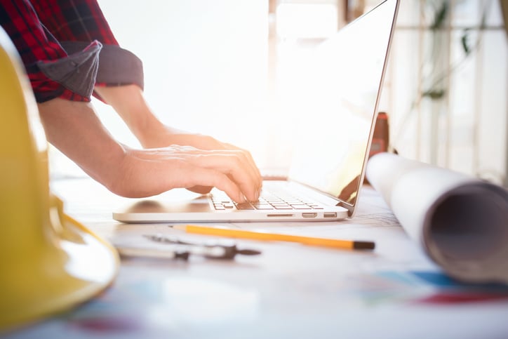 contractor at desk types on laptop beside yellow hard hat