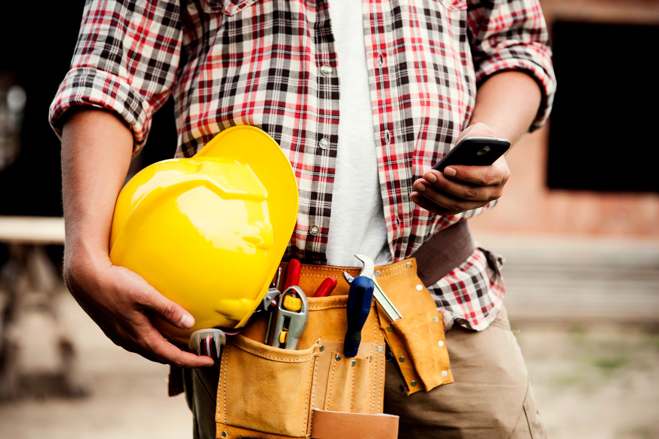 A contractor views smartphone while holding yellow hard hat in right hand