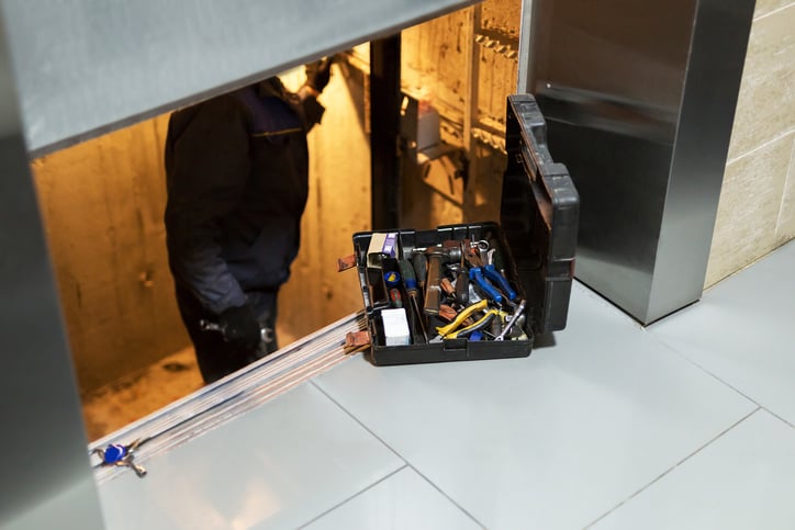 An open toolbox sits on floor beside elevator doorway, slightly ajar showing technician working