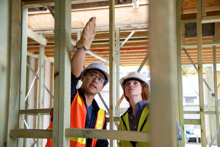 A man and a woman in hard hats examining a lumber frame structure.