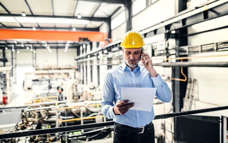 A manufacturing plant manager in hard hat reviews document while speaking on cellphone