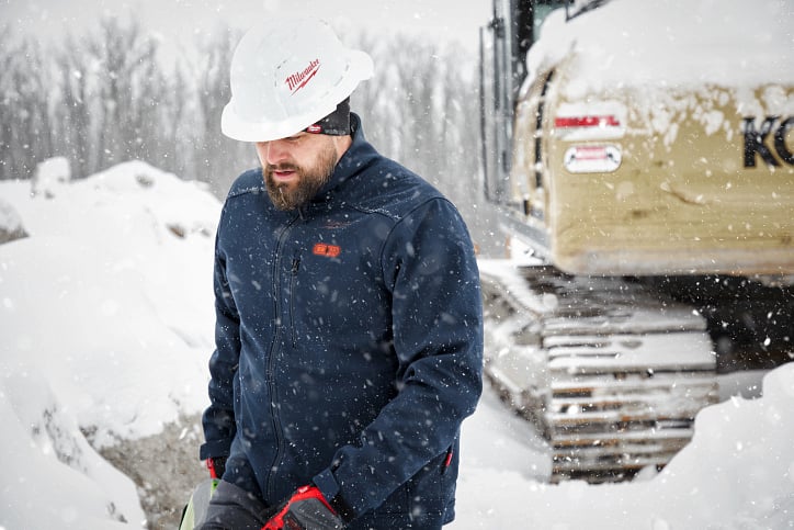 A tradesman in a navy-blue Milwaukee® heated jacket and hard hat walks across snowy jobsite beside excavator 