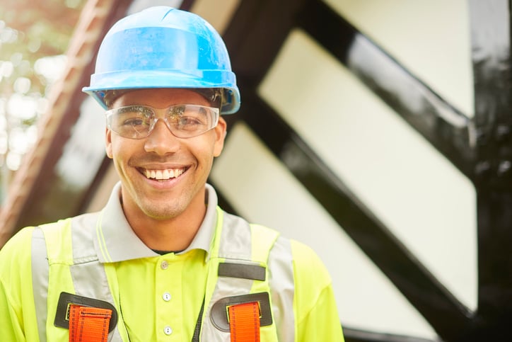 A young roofer in a high-visibility vest, safety glasses, and a blue hardhat