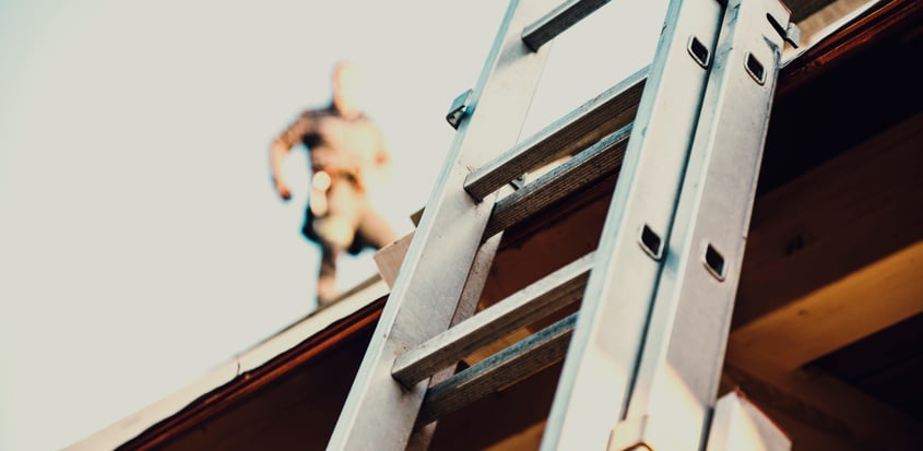 A ladder leans against roof with roofing contractor obscured in the background