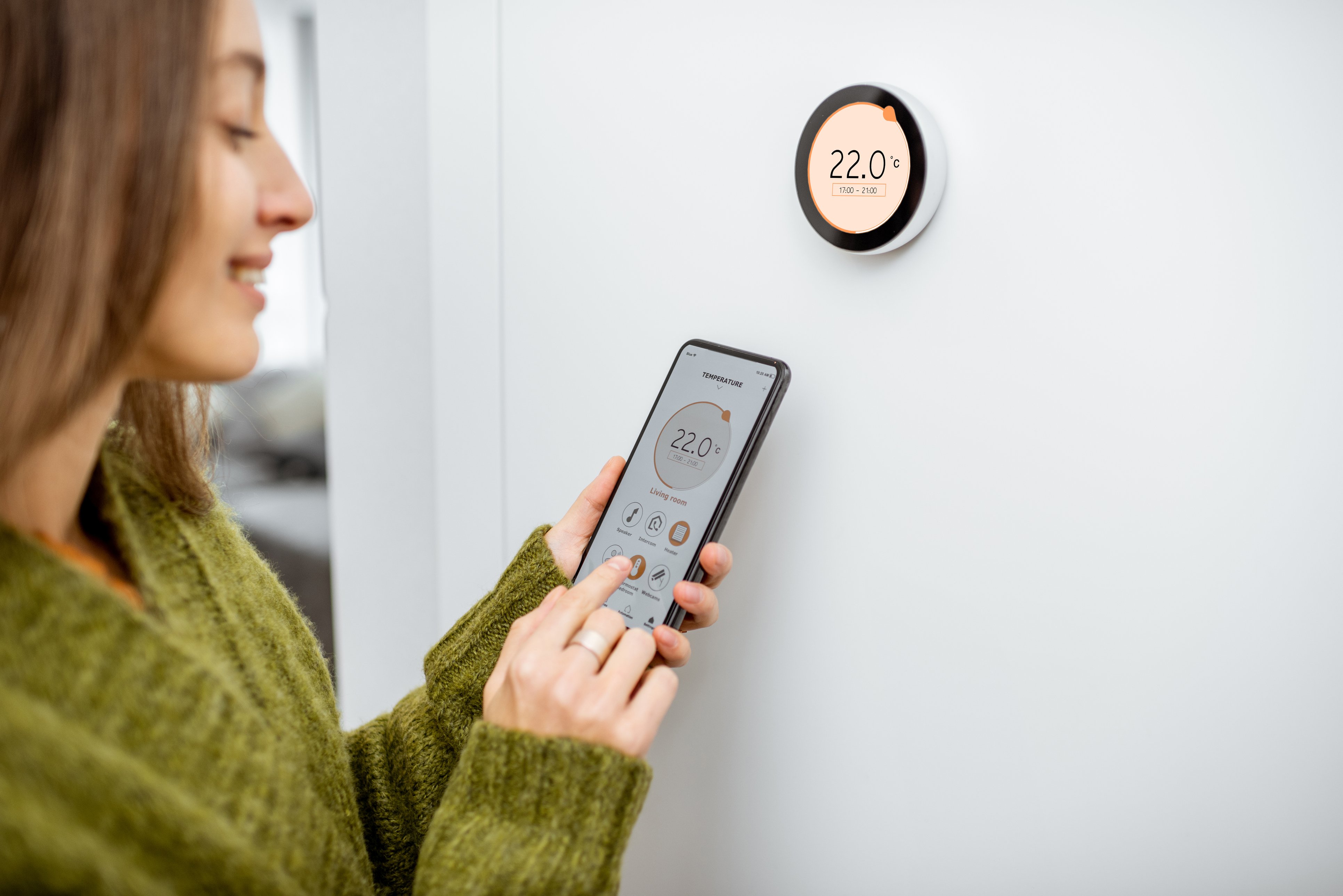 A woman in green sweater uses smart phone beside smart thermostat to adjust temperature