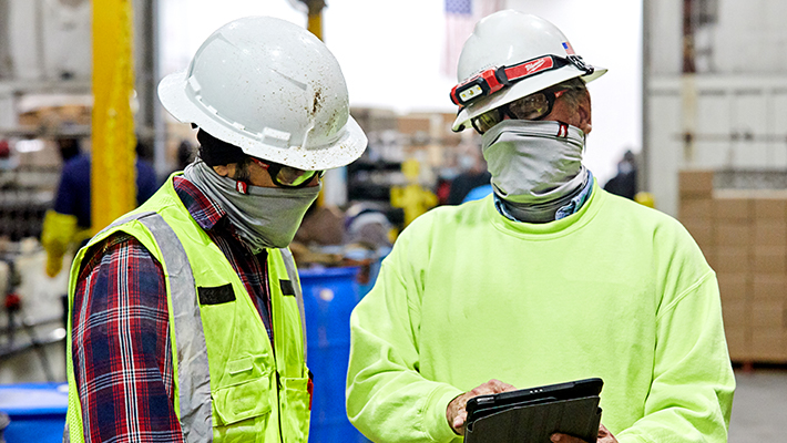 Two warehouse managers wearing gators and hard hats consult inventory over tablet device