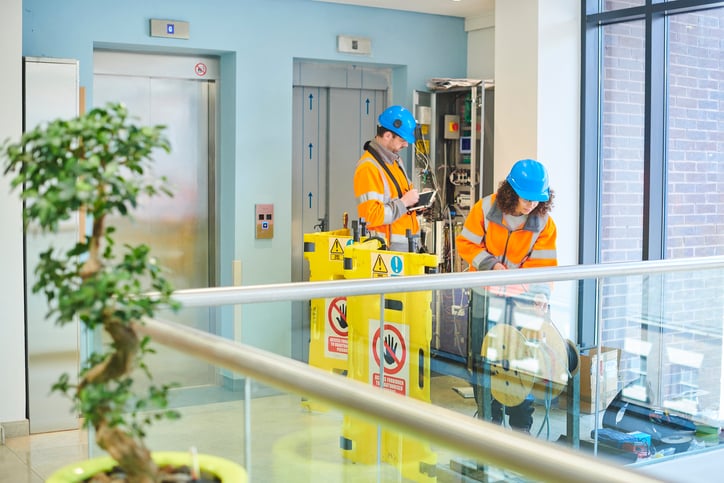 Two elevator mechanics in high-visibility orange jackets service elevator control panel beside safety signs