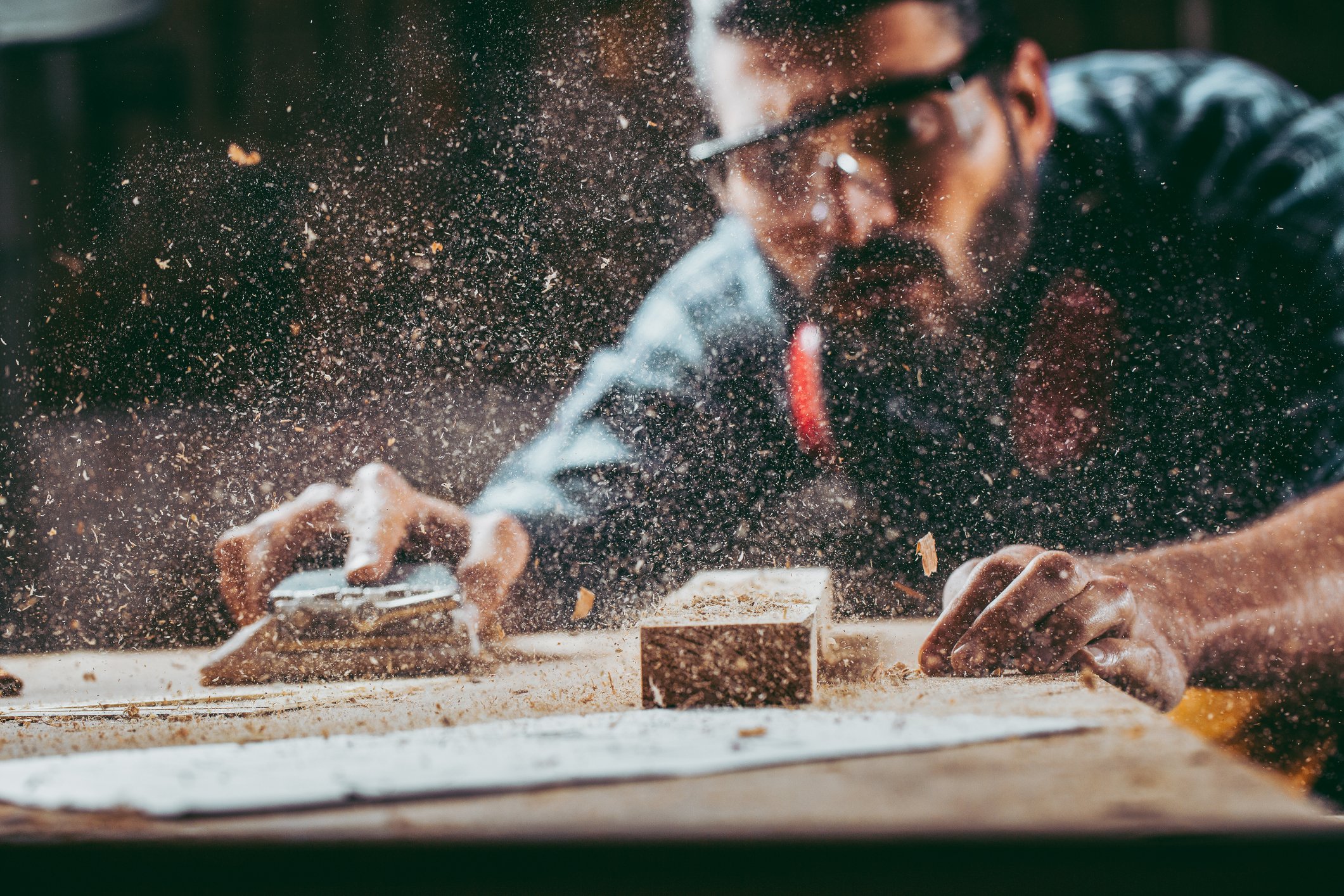 A woodworker wearing safety glasses sands down plywood sheet with sawdust filling the air in front of him